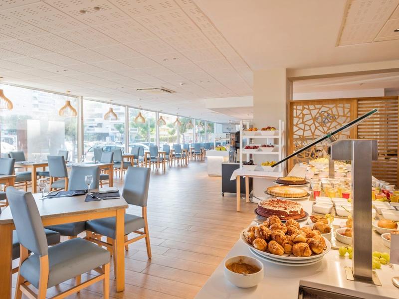 Restaurante luminoso de hotel con mesa buffet llena de pastelería y vista a área con mesas y sillas.
