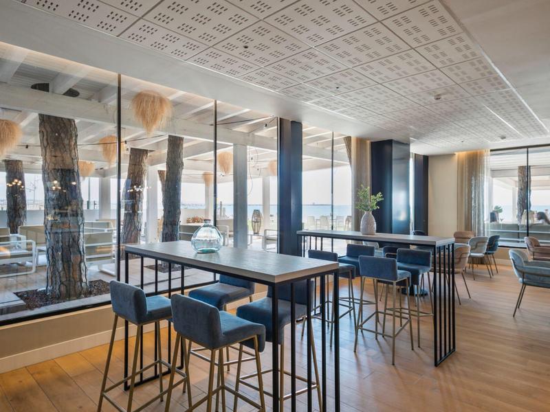 Área de lounge moderna del hotel con mesas altas y sillas, grandes ventanas y acabados en madera clara.