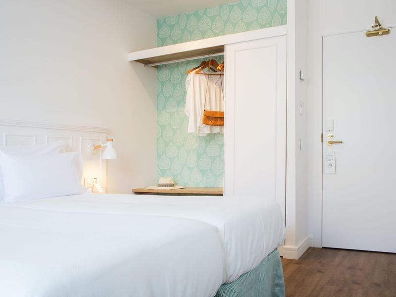 Chambre d'hôtel lumineuse avec lit blanc, mur à motifs et placard ouvert près de la porte.
