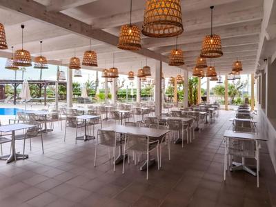 Área de comedor abierta con mesas y sillas blancas y lámparas de mimbre junto a la piscina.