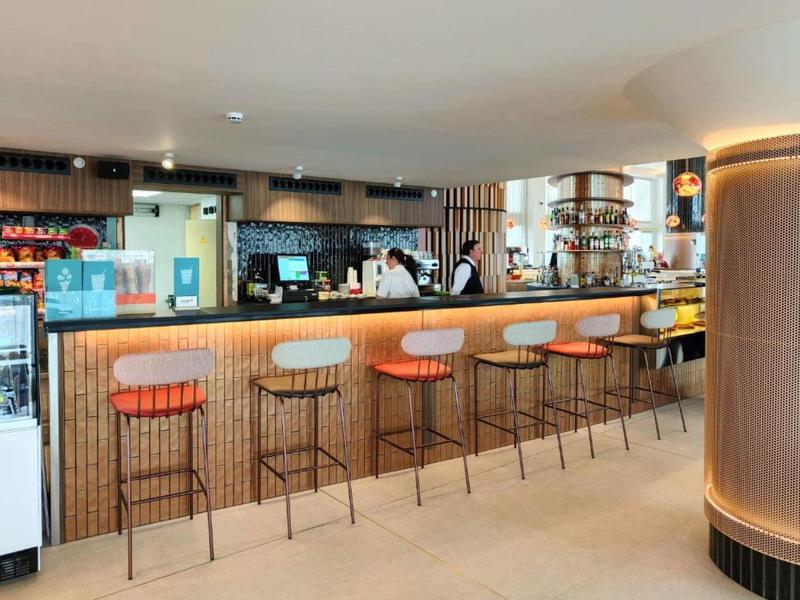 Langer Hotelbar mit moderner Theke und hohen Hockern in warmen Holz- und Orangetönen.