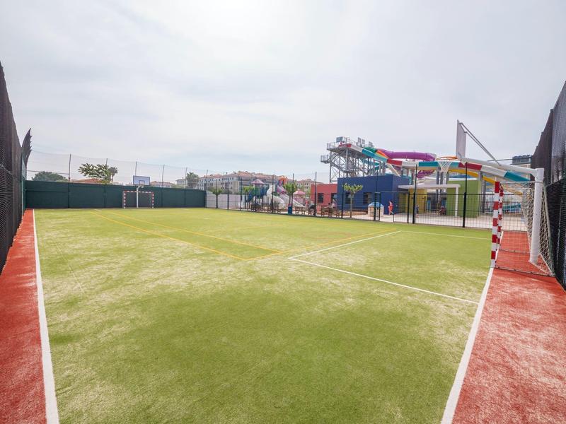 Weitläufiges Fußballfeld mit grünem Kunstrasen und einem angrenzenden Spielplatz mit farbenfrohen Rutschen.