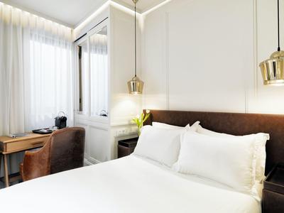 Světý hotelový pokoj s bílou postelí, hnědým křeslem, dřevěným stolem a moderním osvětlením.