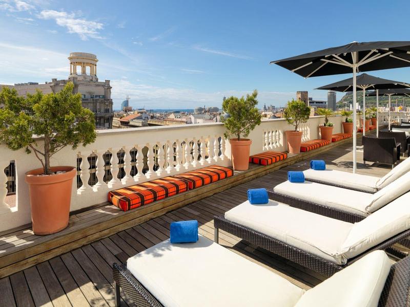 Slunná střešní terasa s lehátky, slunečníky, květináči a výhledem na město.