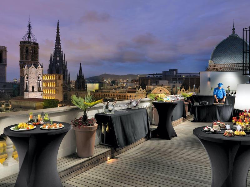 Venkovní střešní terasa s koktejlovými stolky a městskou krajinou za soumraku.