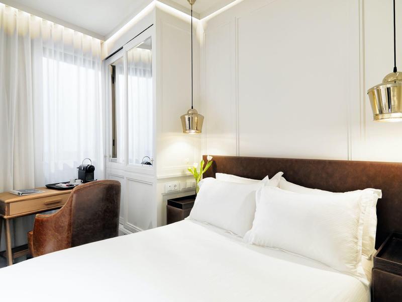 Světý hotelový pokoj s bílou postelí, hnědým křeslem, dřevěným stolem a moderním osvětlením.