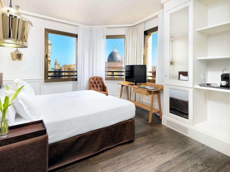 Světle hotelový pokoj s velkou postelí, výhledem na město, stolem a moderními bílými policemi.