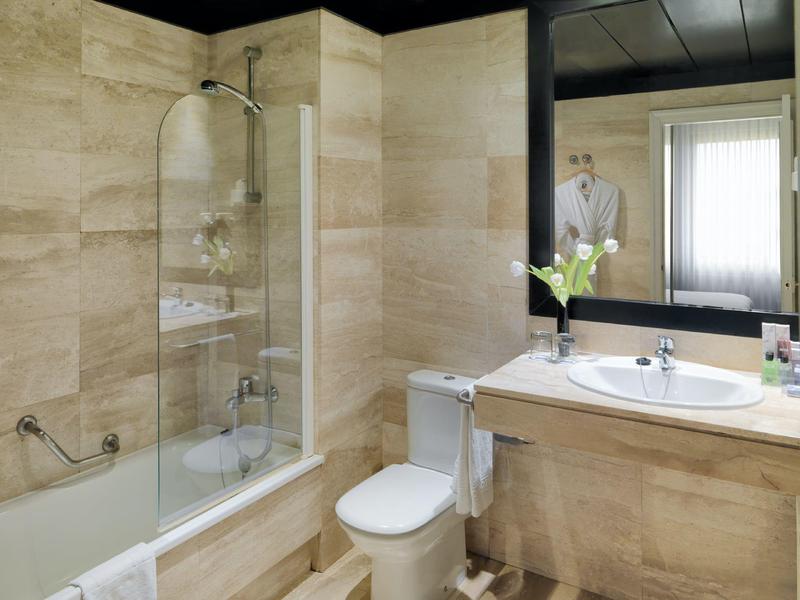 Moderní hotelová koupelna s béžovými dlaždicemi, vanou, toaletou, velkým zrcadlem a umyvadlem.