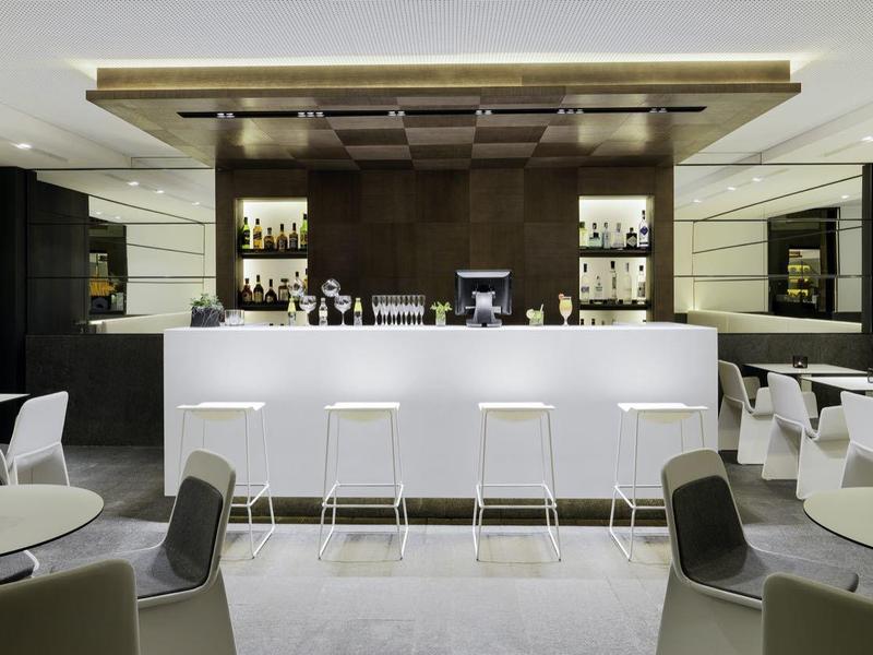 Moderner Barbereich mit weißer Theke, hohen Hockern und stilvollen grauen Sesseln in hellem Ambiente.