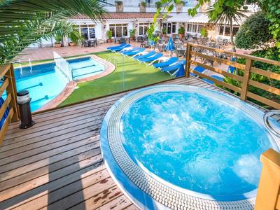 Whirlpool mit sprudelndem Wasser auf Holzdeck neben Pool und blauen Liegen in Gartenanlage.