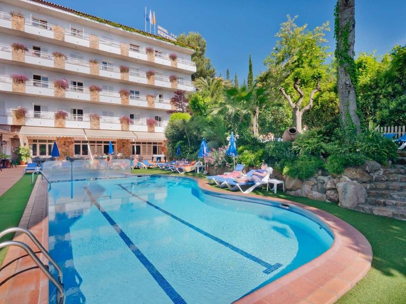 Hotel met groot zwembad, ligstoelen en tuin onder blauwe lucht.