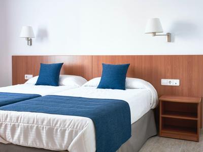 Moderní hotelový pokoj s manželskou postelí, modrými polštáři a přehozem, jednoduchým nočním stolkem a nástěnnými lampami.