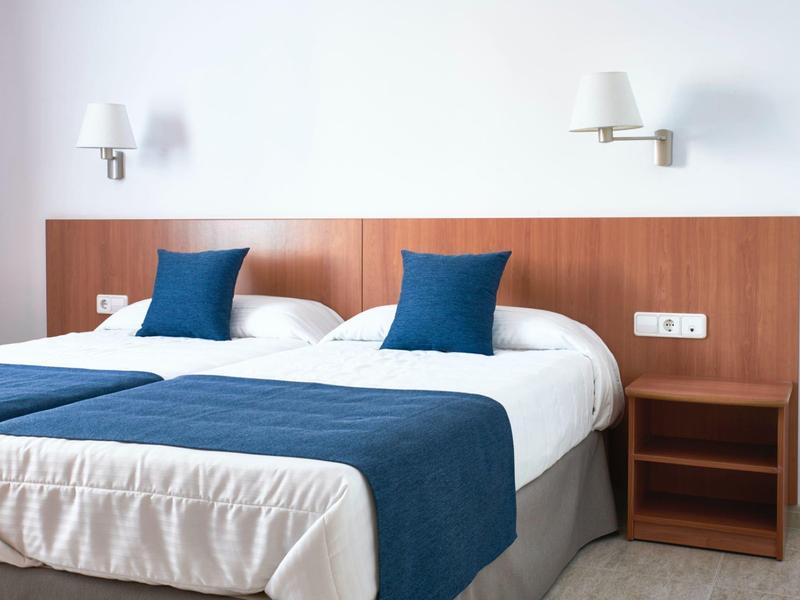 Moderní hotelový pokoj s manželskou postelí, modrými polštáři a přehozem, jednoduchým nočním stolkem a nástěnnými lampami.