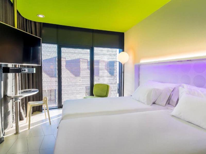 Modernes Hotelzimmer mit grün-gelber Decke, zwei weißen Betten und großen Fenstern zum Balkon.