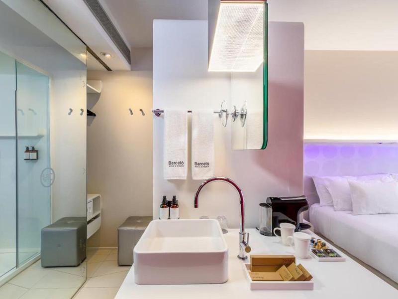 Modernes Hotelzimmer mit weißem Waschbecken, Glasdusche und lila beleuchtetem Bett.