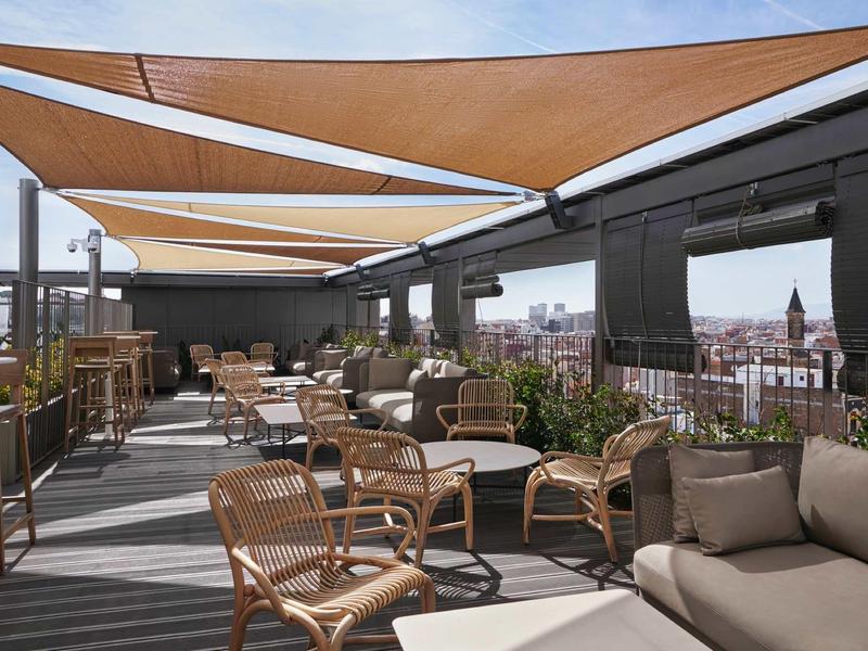 Moderní střešní terasa s pohovkami a proutěnými židlemi pod stínícími plachtami.