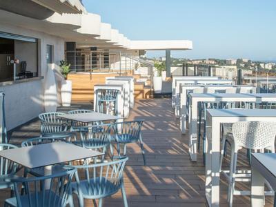 Terrasse mit weißen Tischen und grauen Stühlen mit Meerblick unter klarem Himmel.