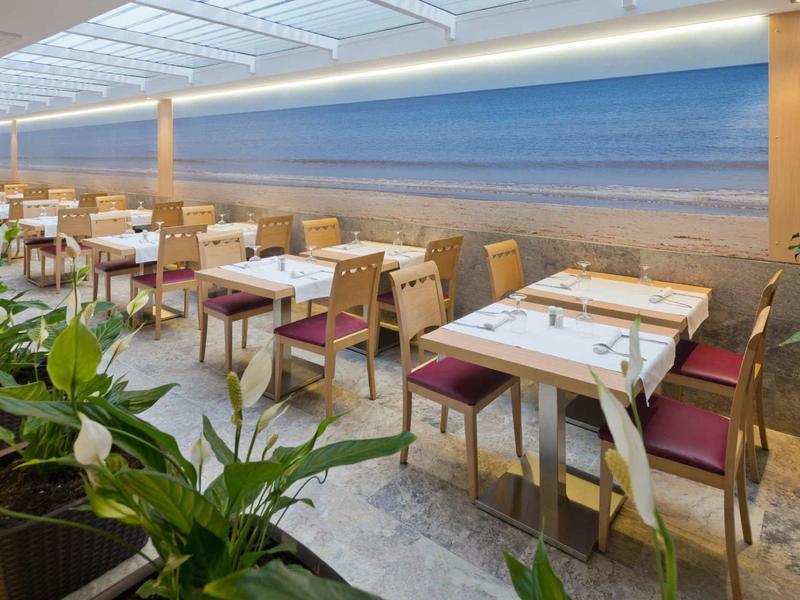 Modernes Restaurant mit Holztischen, Stühlen mit roten Polstern und Wandbild eines Strandes.