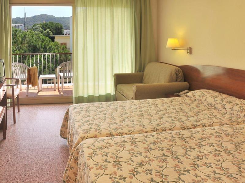 Hotelzimmer mit zwei Einzelbetten, Blumenbettwäsche, Sesseln, Fernseher und Balkon mit grünem Ausblick.