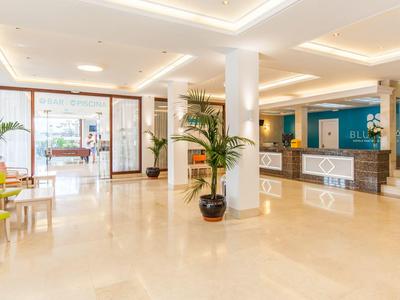 Jasna lobby hotelowa z recepcją, roślinami i częścią wypoczynkową z dużymi oknami.