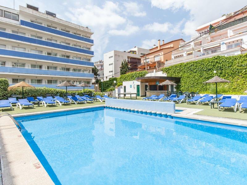 Duży basen na zewnątrz z leżakami między nowoczesnymi budynkami hotelowymi pod błękitnym niebem.