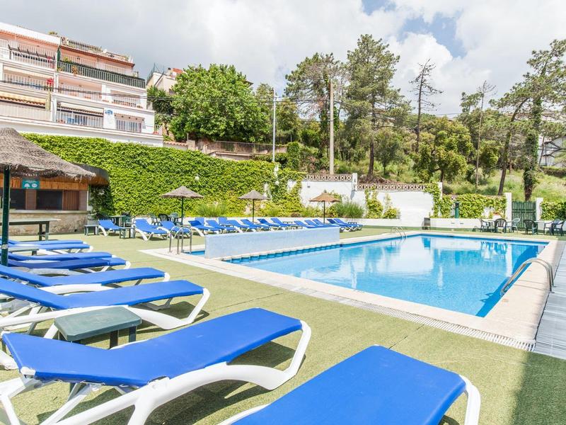 Strefa basenowa z niebieskimi leżakami i zielonymi roślinami wokół hotelu.