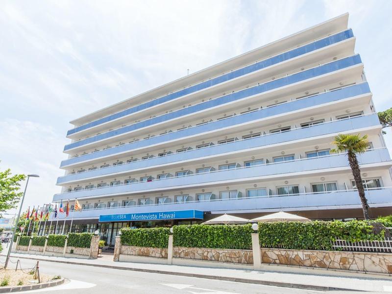 Nowoczesny budynek hotelu z wieloma balkonami i niebieskimi markizami pod zachmurzonym niebem.