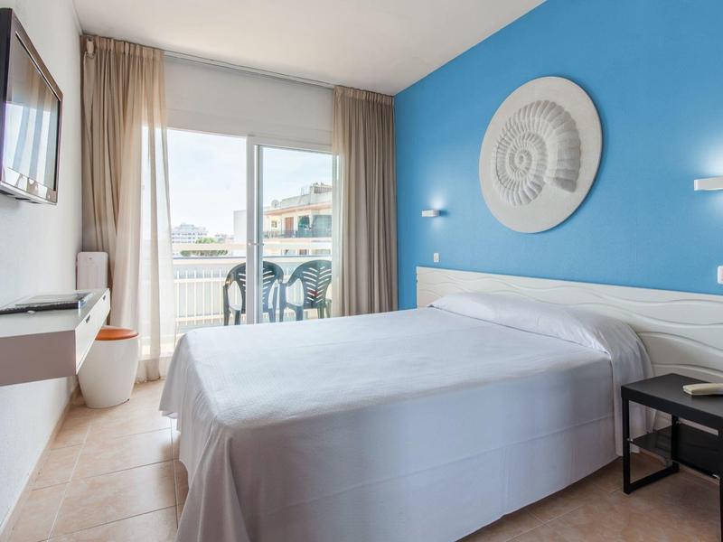 Jasny pokój hotelowy z podwójnym łóżkiem, balkonem i niebieską ścianą akcentową z dekoracją muszli.