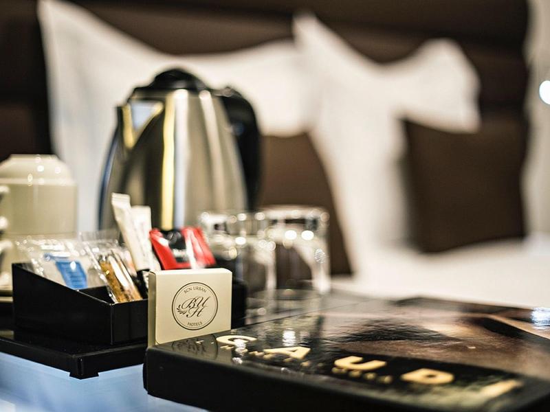 Kaffeekocher, Tassen und Zucker auf einem Tisch vor einem Bett mit weißen Kissen und dunkler Decke.