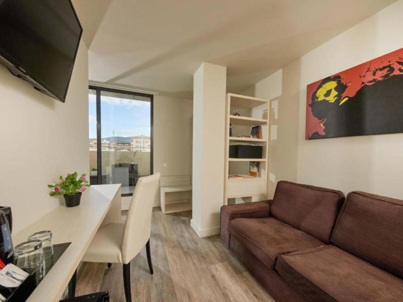 Kleines Zimmer mit braunem Sofa, weißem Tisch, Stühlen, Fernseher und buntem Wandbild.