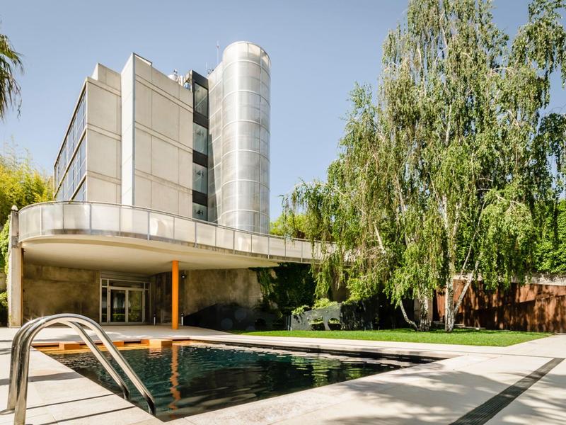 Modern hotel pool with metal ladder surrounded by tall trees and buildings.