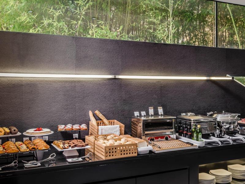 Buffet with assorted pastries, bread, and drinks against dark wall and window.