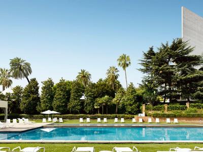 Moderner Hotelpool mit Liegestühlen, Palmen und klar blauem Himmel im Hintergrund.