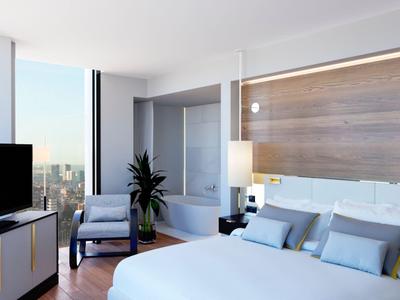 Modern hotel room with large bed, TV, armchair, and city view.