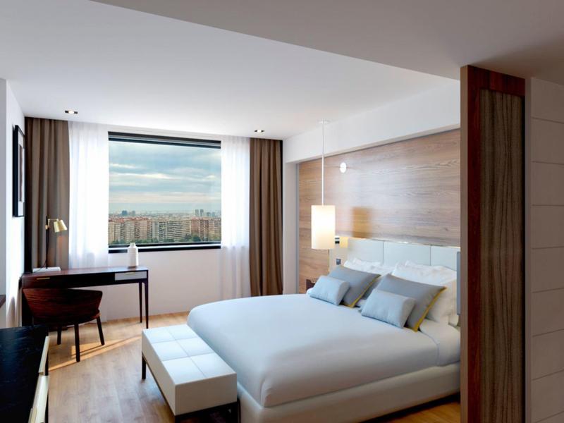 Modern hotel room with large bed, desk, TV, and large window with city view.
