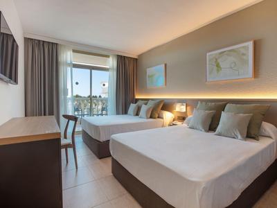 Světle hotelový pokoj se dvěma postelemi, pracovním stolem, židlí a velkým oknem se záclonami.