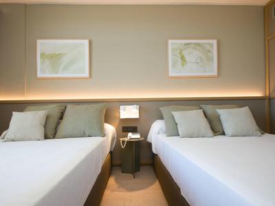 Hotelový pokoj se dvěma velkými manželskými postelemi, šedými stěnami a nepřímým osvětlením.