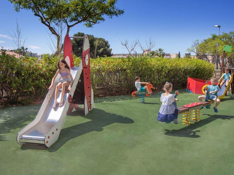 Hřiště s klouzačkou a houpačkou, děti si hrají pod stromy a modrou oblohou.