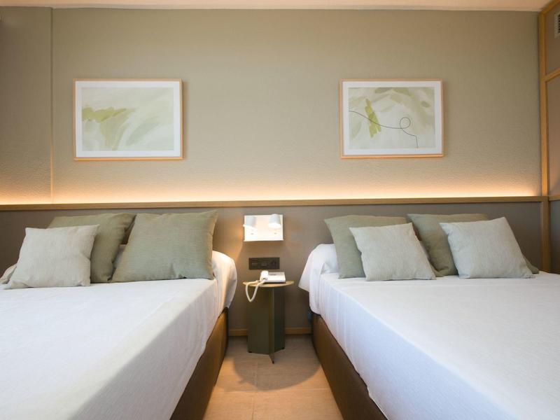 Hotelový pokoj se dvěma velkými manželskými postelemi, šedými stěnami a nepřímým osvětlením.