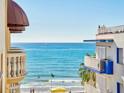 Meerblick zwischen weißen Häusern mit Balkonen, blauer Himmel, Sandstrand und Menschen am Wasser.