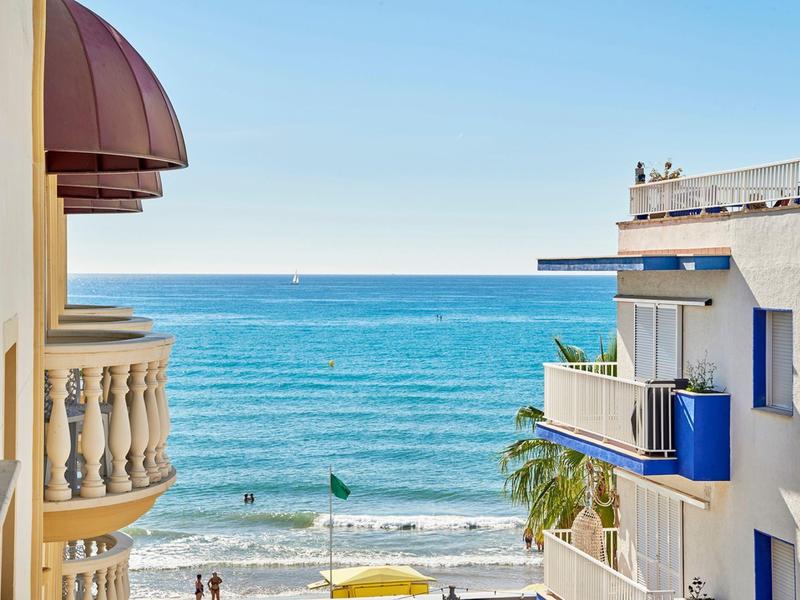 Blauwe lucht en zeezicht tussen gebouwen met balkons en mensen op het strand.