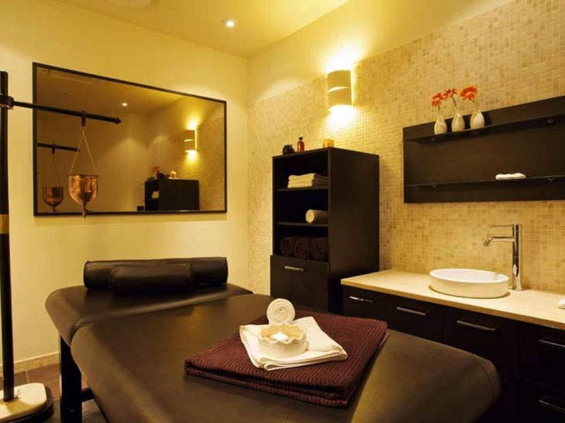 Behandlungsraum mit dunkler Massageliege, Handtüchern, Spiegel und warmen, gelben Wänden.