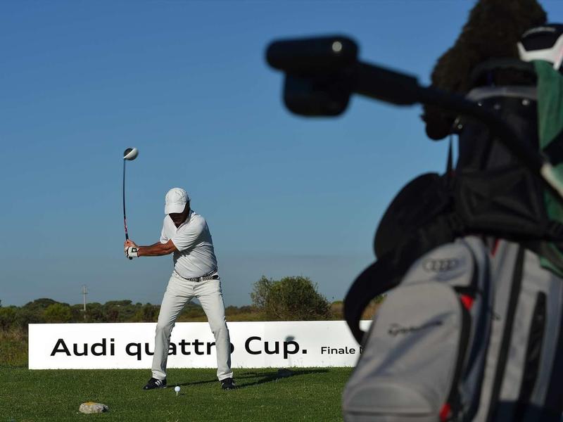Golfer in weißer Kleidung beim Abschlag auf grüner Wiese, blauer Himmel, Audi Quattro Cup Schild.