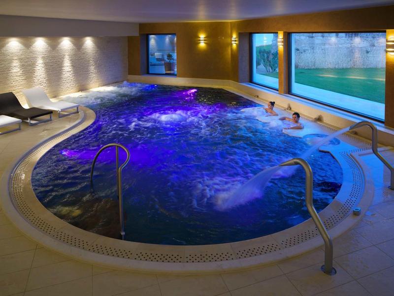 Innenraum mit rundem Whirlpool, blau beleuchtet, mit Liegestühlen und großen Fenstern.