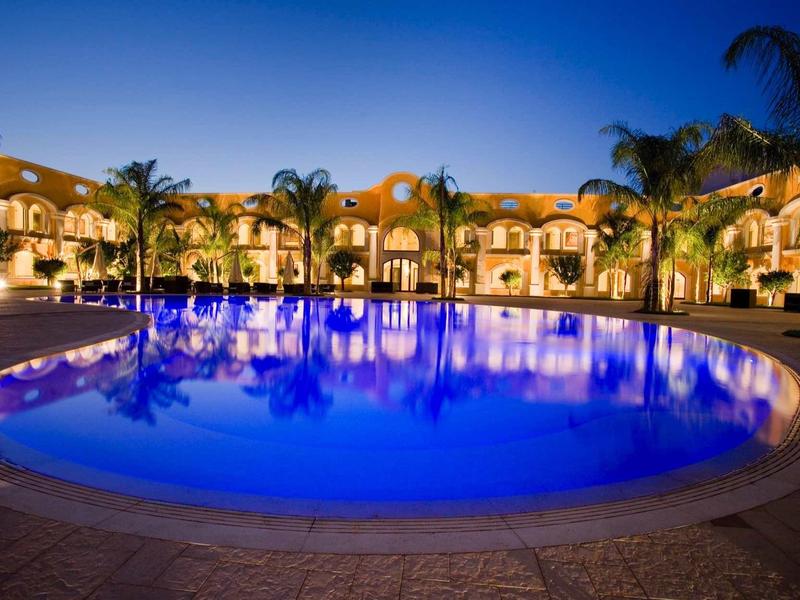 Rundes Schwimmbecken mit blauem Wasser, umgeben von Palmen und beleuchteten Gebäuden bei Nacht.