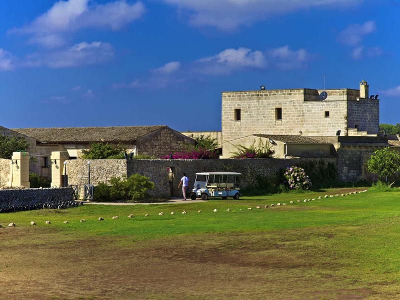 Weißes, rechteckiges Gebäude auf grünem Feld unter blauem Himmel mit vereinzelten Wolken.