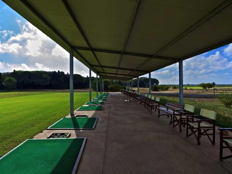 Überdachter Golf-Übungsbereich mit Abschlagmatten, Bänken und grünem Rasen unter blauem Himmel.