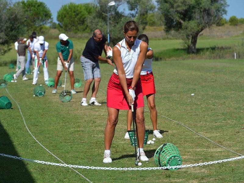 Mehrere Personen üben Golf auf einer Grünfläche, vorne eine Frau mit Golf-Driver in rotem Rock.