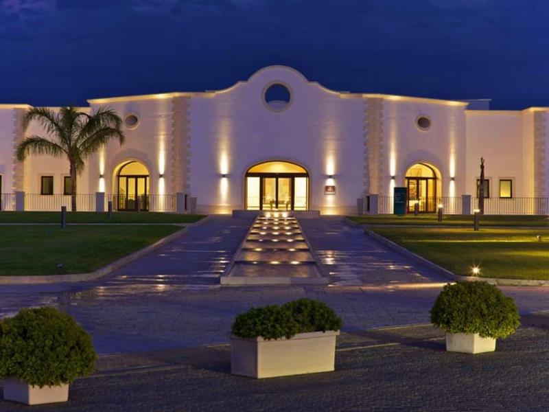 Hell beleuchtetes, weißes Gebäude mit Säulen, Pool, Palmen und Buschkübeln bei Nacht.