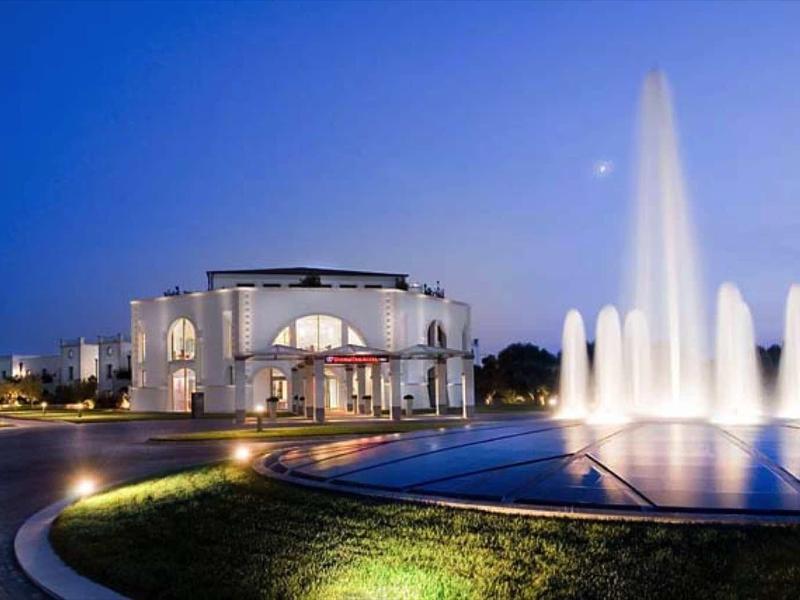Beleuchteter Springbrunnen mit hohem Wasserstrahl vor einem weißen Gebäude bei Abenddämmerung.